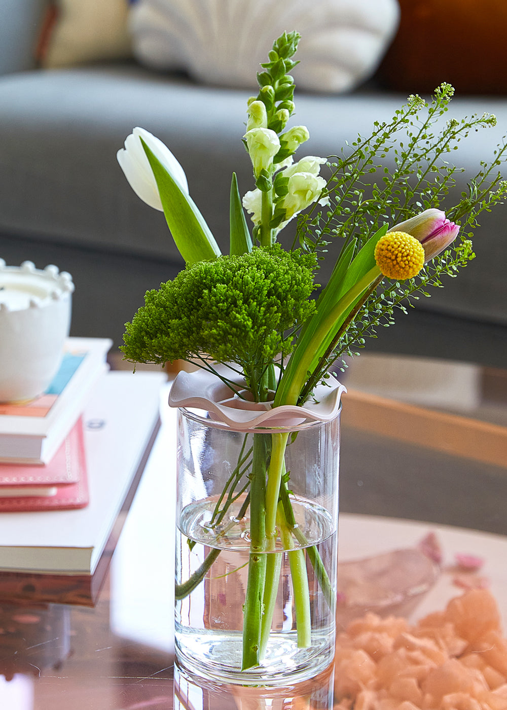 Clear glass vase with fresh white tulips, yellow and pink flowers, and green foliage, set on a stylish coffee table.
