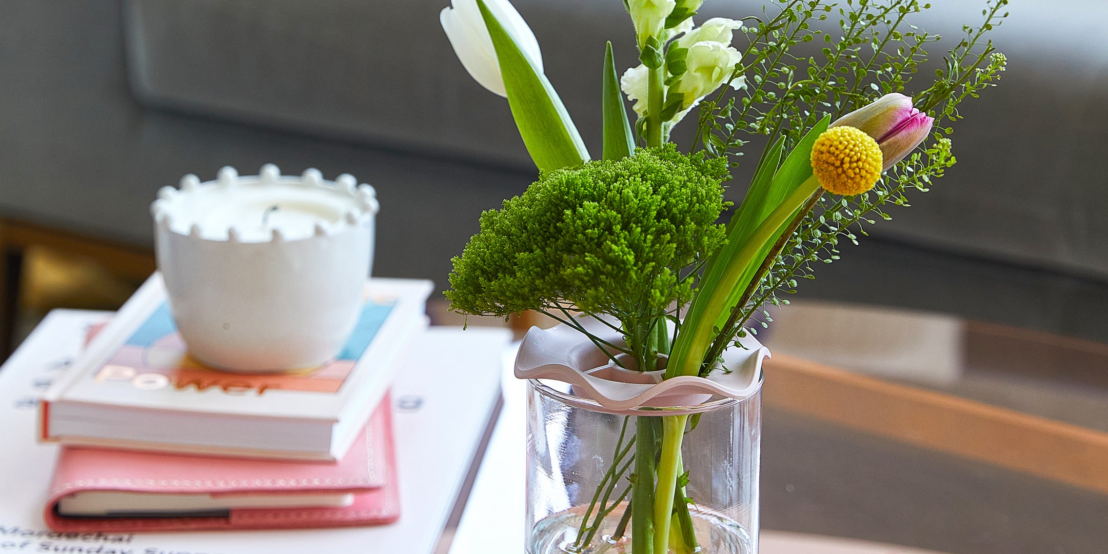 Vibrant indoor garden with fresh green herbs and colorful flowers in a clear glass vase, complemented by a candle and books.
