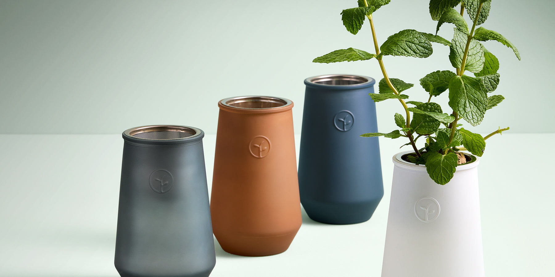 Four stylish hydroponic pots in matte finishes: gray, terracotta, navy, and white, with a mint plant in the white pot.