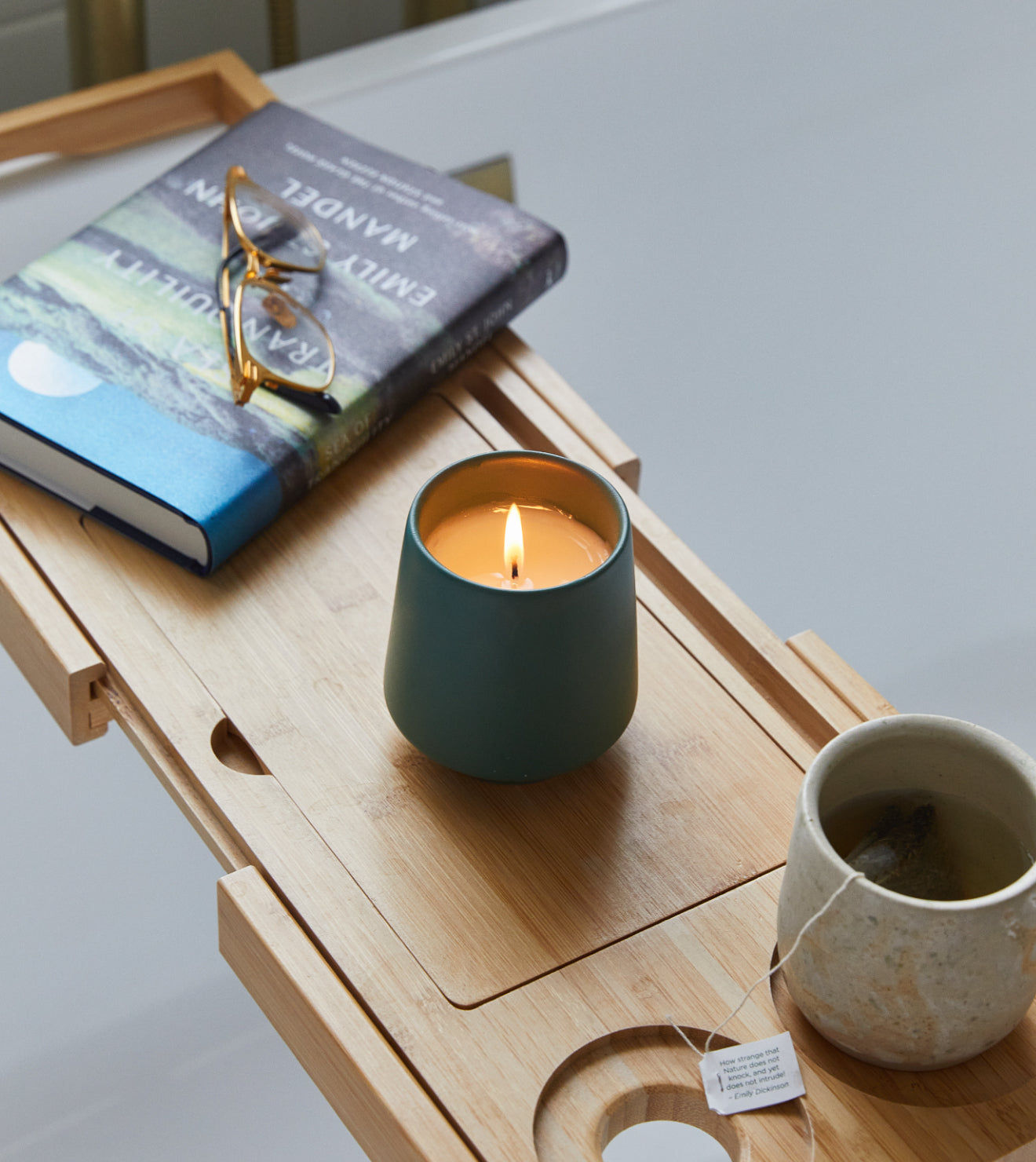 A bamboo indoor garden tray with a lit teal candle, a beige ceramic mug, a book, and sunglasses on top.