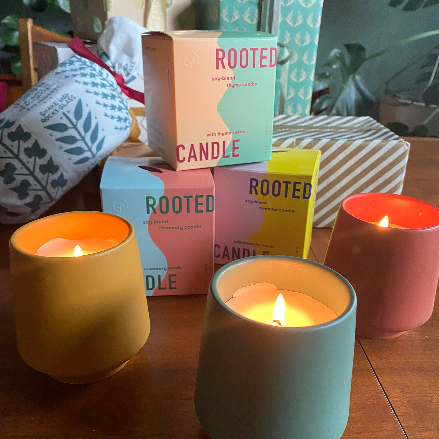 Colorful soy-blend candles in yellow, pink, and blue containers with lit wicks, alongside pastel gift boxes in pink, blue,...