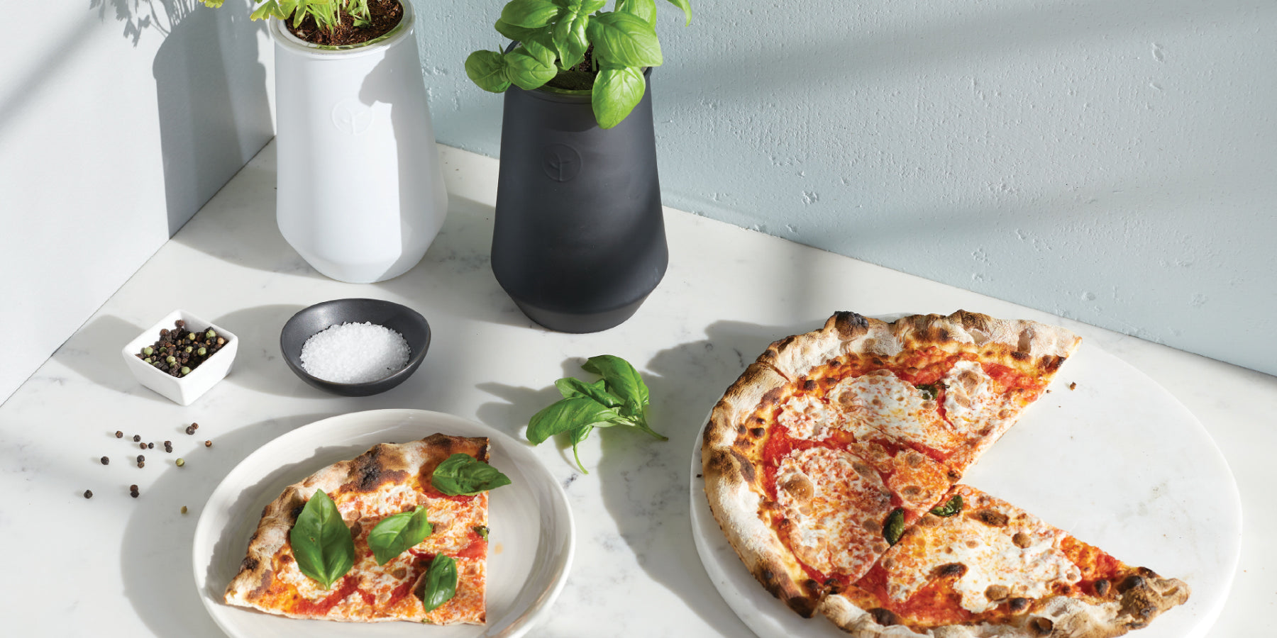 Indoor hydroponic herb garden with basil and lettuce in black and white planter pots, next to a pizza on white marble surf...