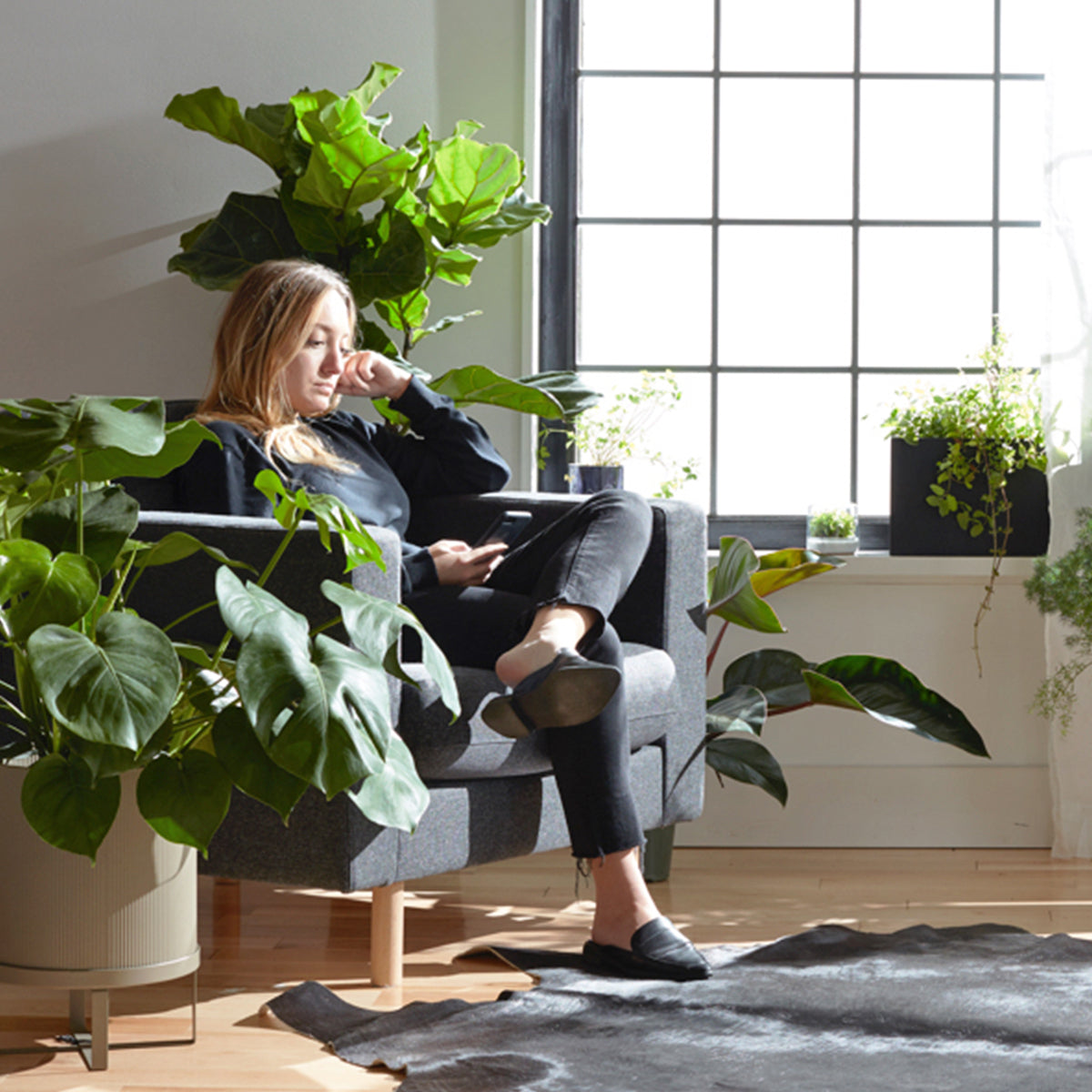 Woman relaxing in a chair surrounded by lush indoor plants in a sunlit room.