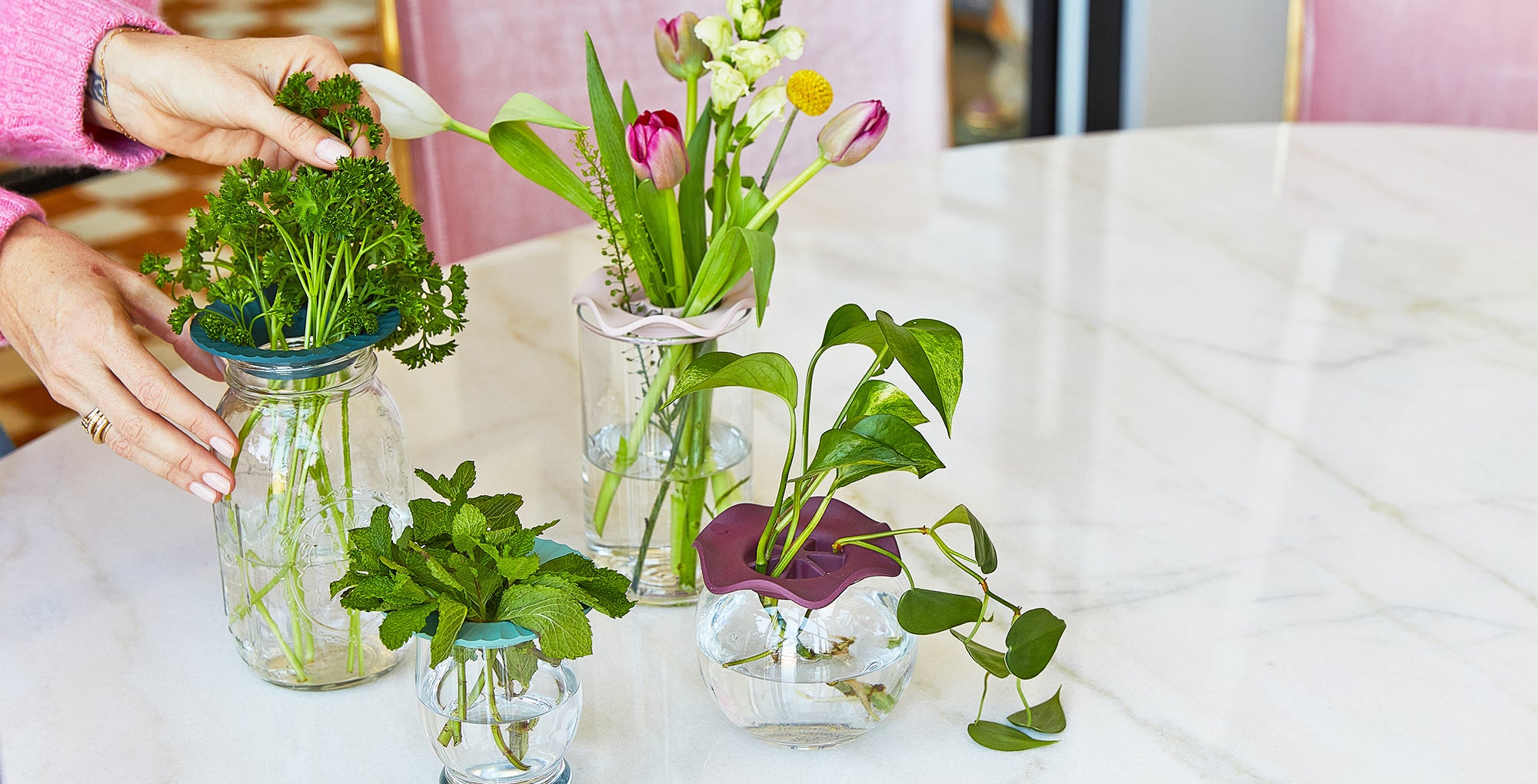 Hand placing fresh herbs and flowers in stylish glass jars with silicone lids on a marble table, showcasing vibrant greens...