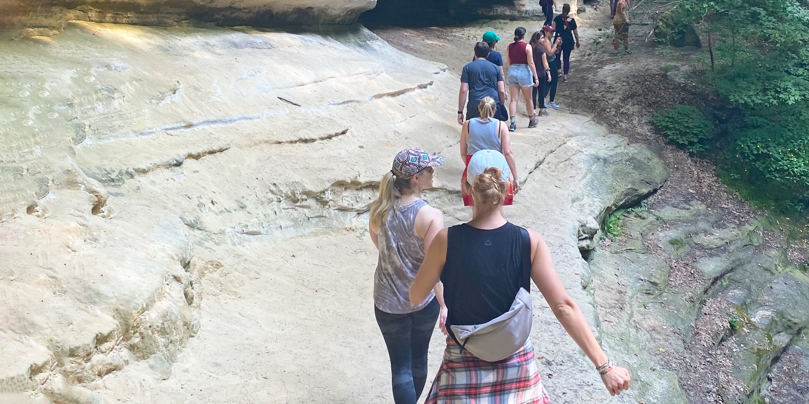 A group of people hiking on a rocky path through a lush green forest, wearing casual outdoor clothing and hats.