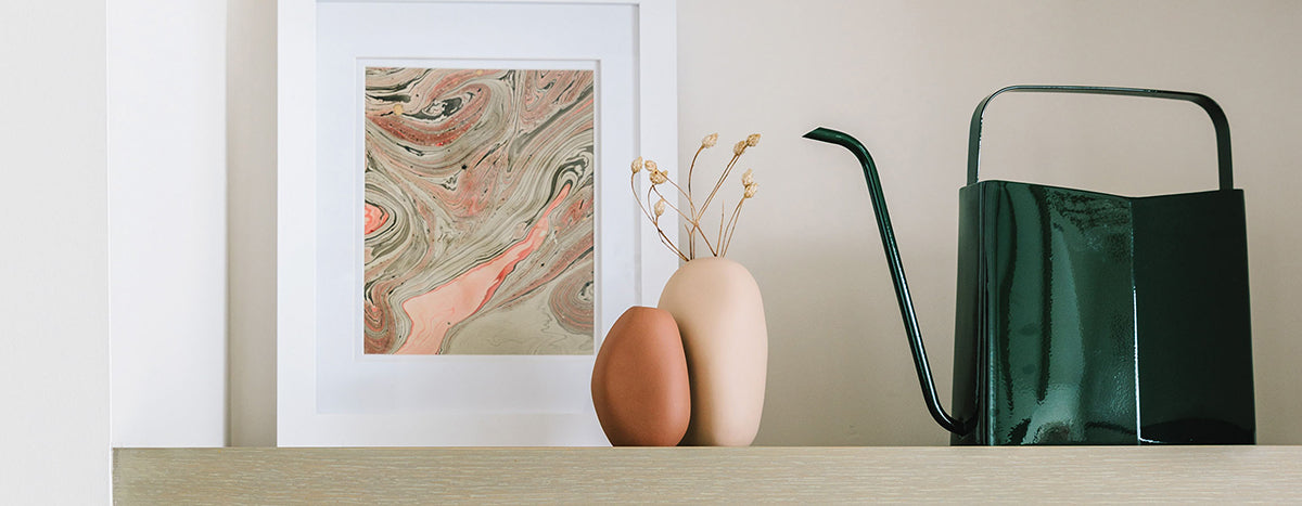 Stylish indoor display featuring a green watering can, two decorative vases in neutral tones, and abstract wall art.
