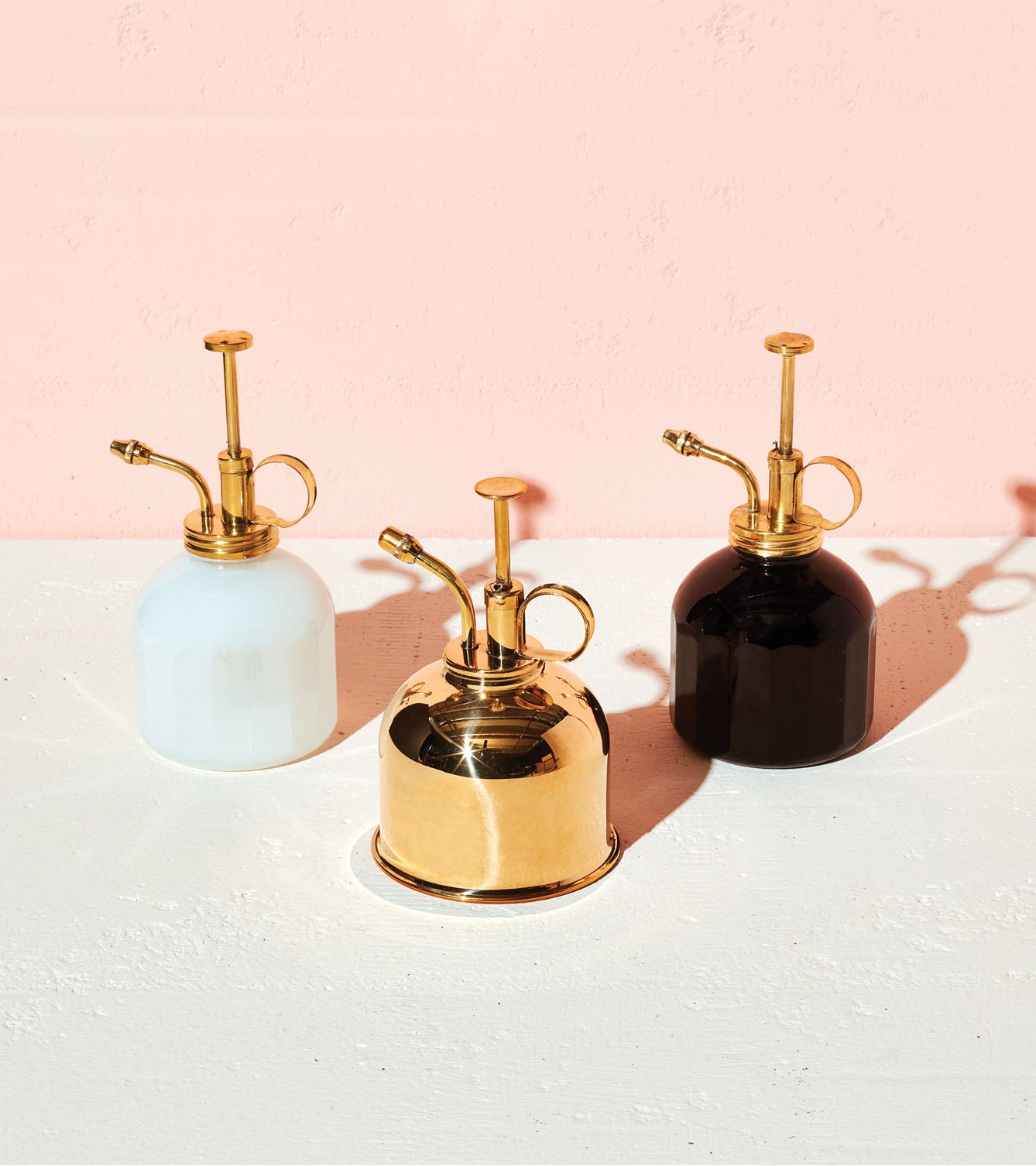 Three stylish plant misters in white, black, and gold, featuring elegant brass nozzles against a pastel pink background.