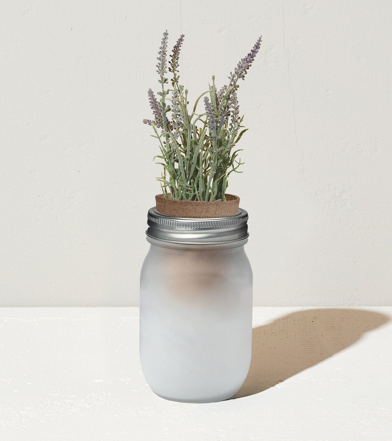 Frosted glass jar with a metal lid, featuring lavender plants and a cork top, set against a neutral background.
