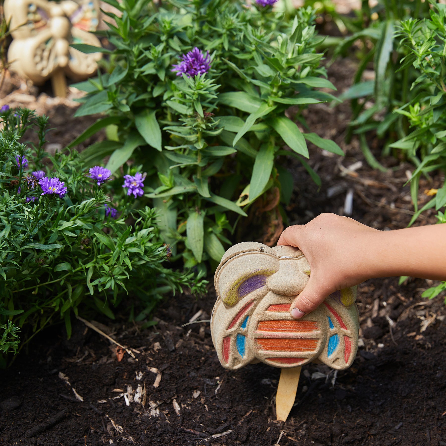 A child's hand holds a colorful garden marker while planting in a flower bed with purple blooms and green foliage.