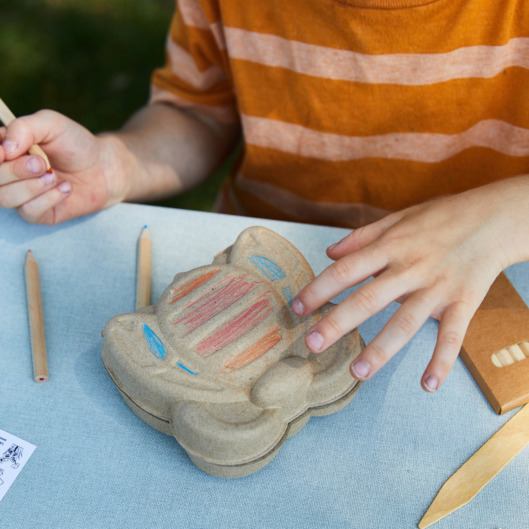Child decorating a cardboard garden kit shaped like a critter, using colored pencils on a light blue table.