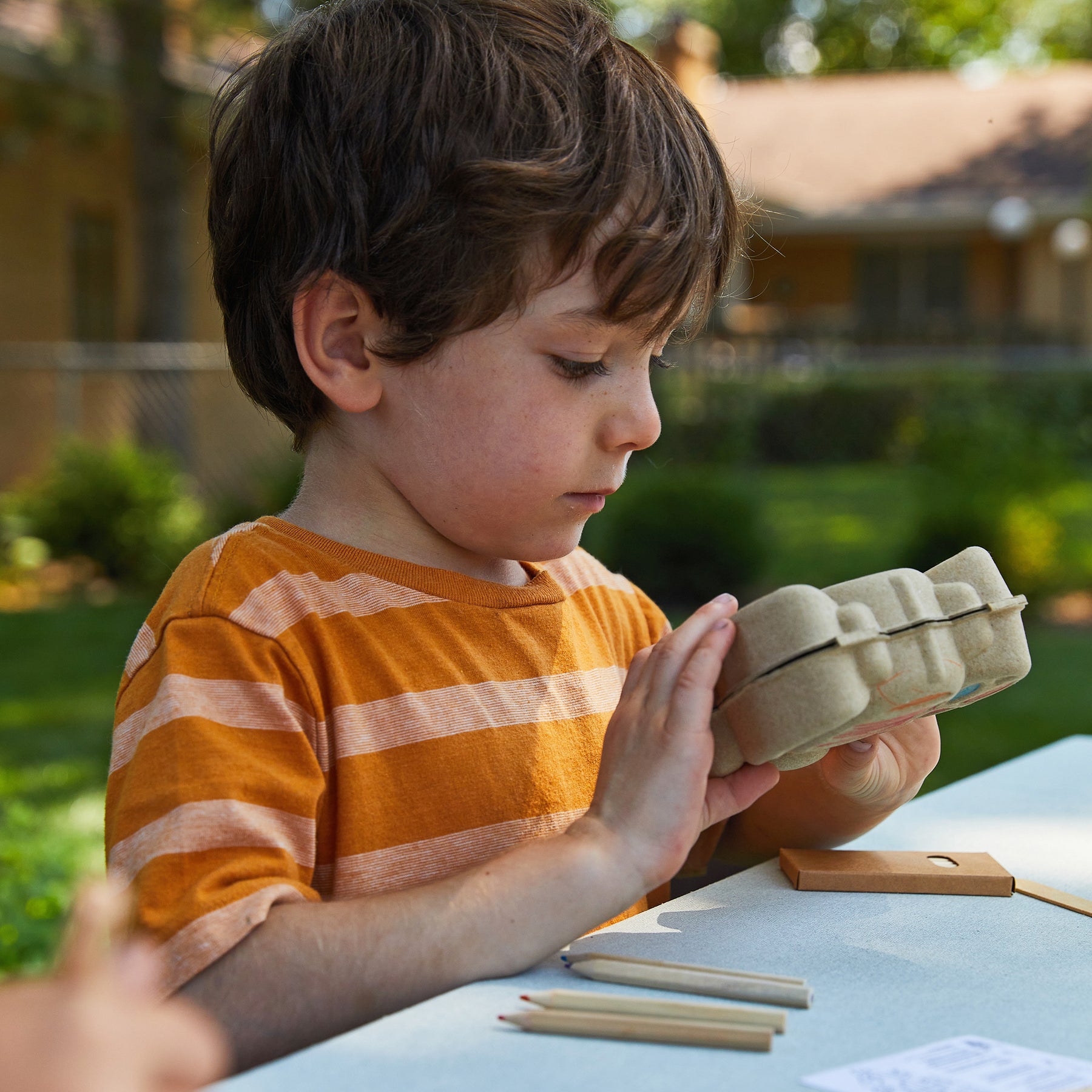 A young boy examines a Curious Critters Garden Kit while sitting at a table outdoors, surrounded by greenery.