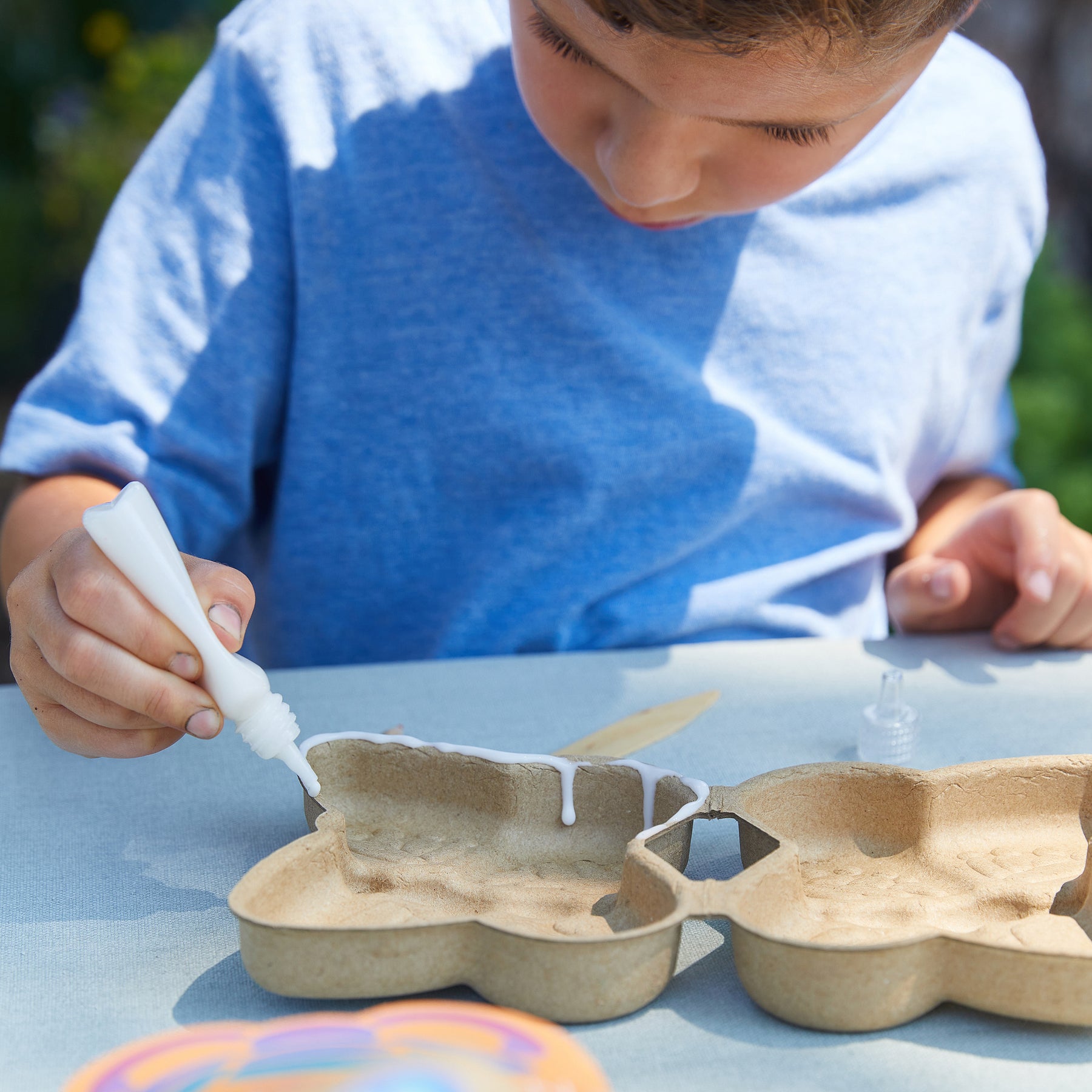 A child applies glue to a garden kit mold while focused on a crafting project outdoors.