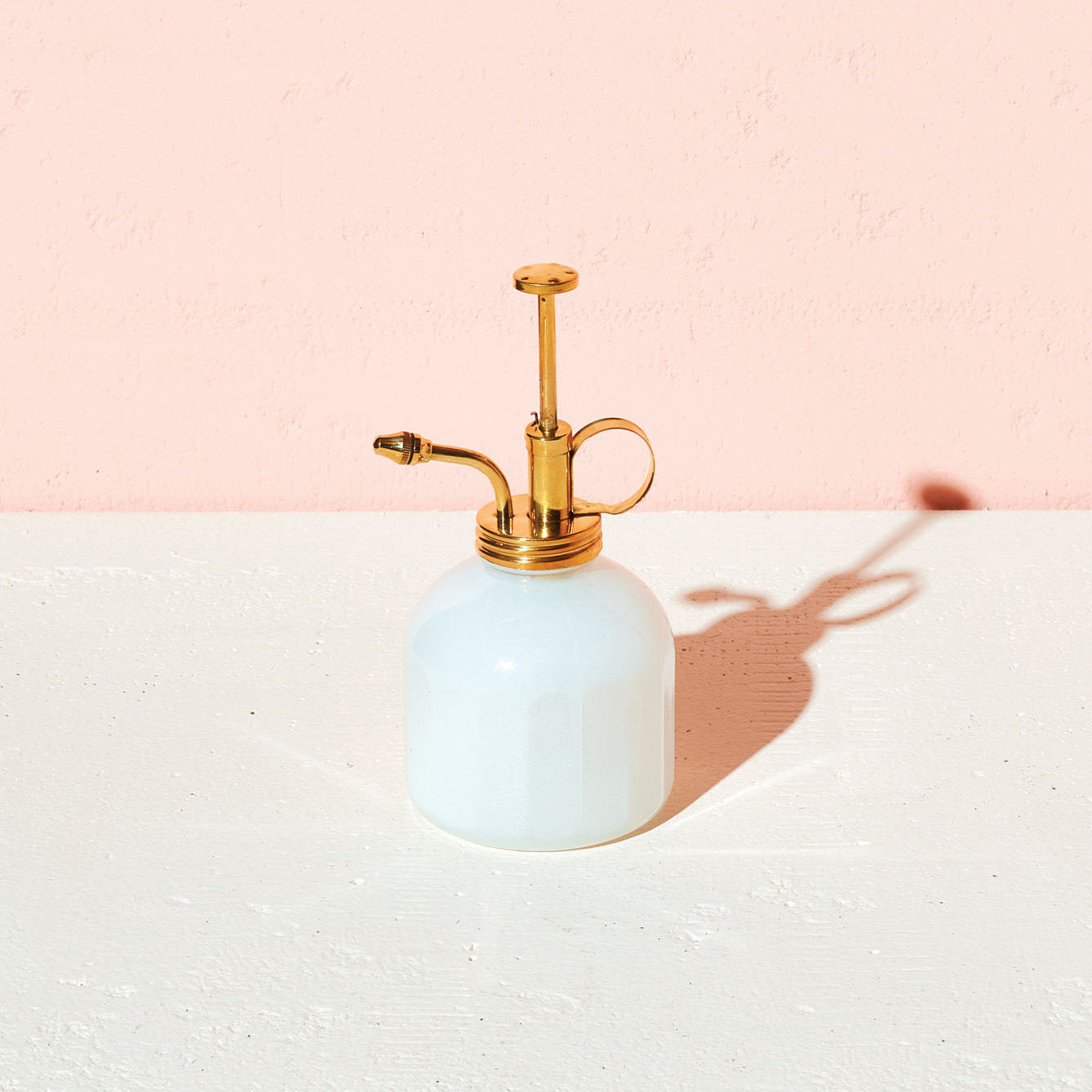 Elegant brass and white glass plant mister on a light surface, casting a soft shadow against a pink background.