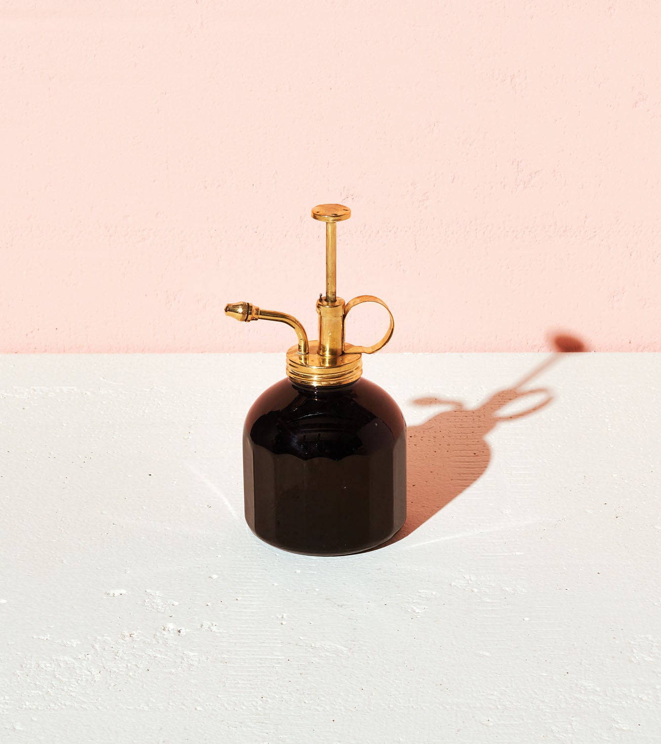 Black glass mister with a brass nozzle and handle, casting a shadow on a white surface against a pink background.