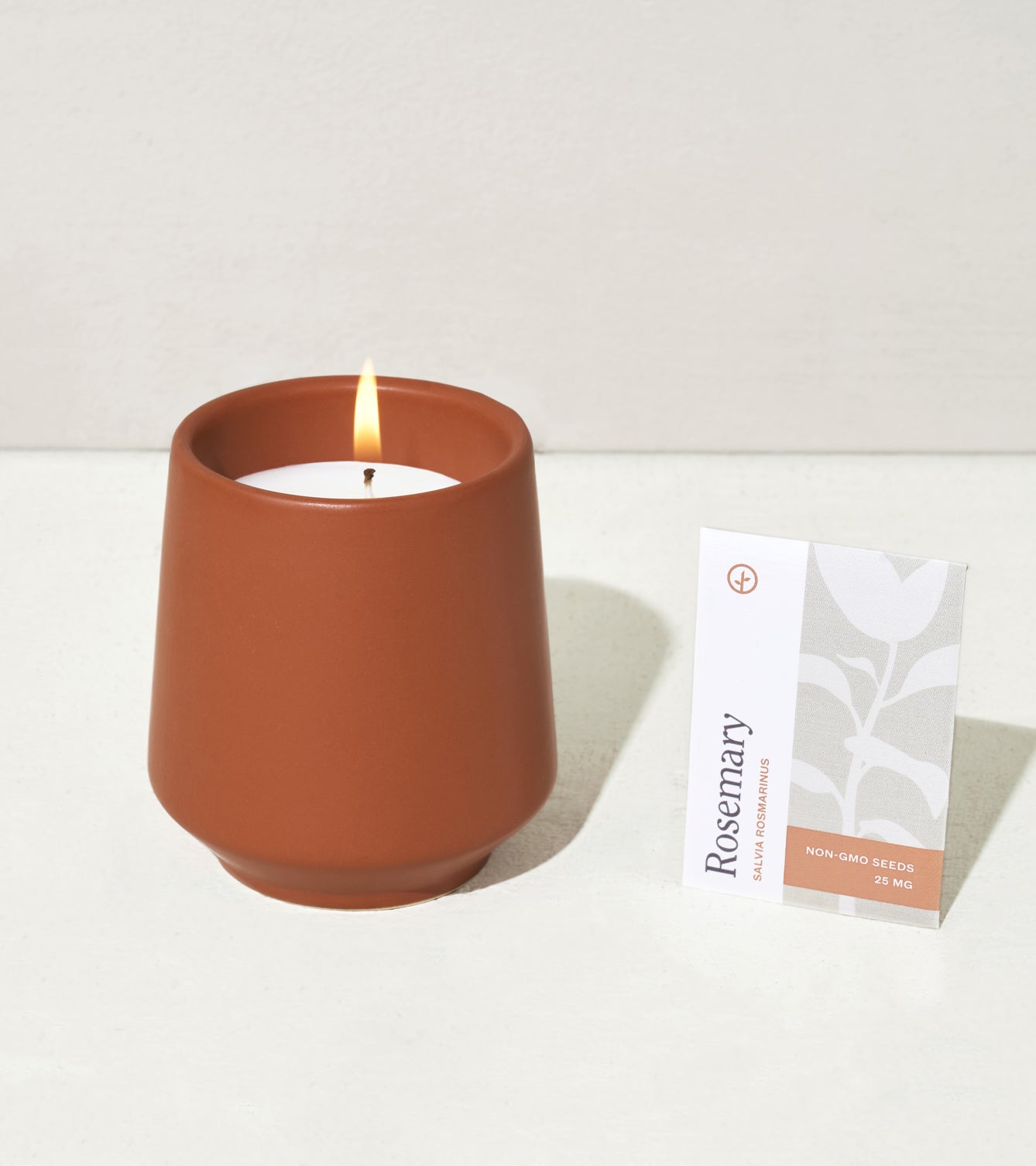 A terracotta candle holder with a lit candle beside a packet of rosemary seeds labeled as non-GMO.