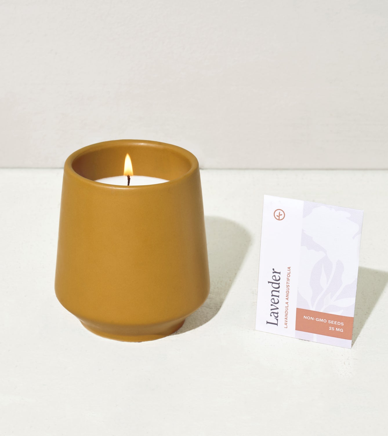 A lavender-scented candle in a mustard-colored ceramic holder next to a packet of non-GMO lavender seeds.