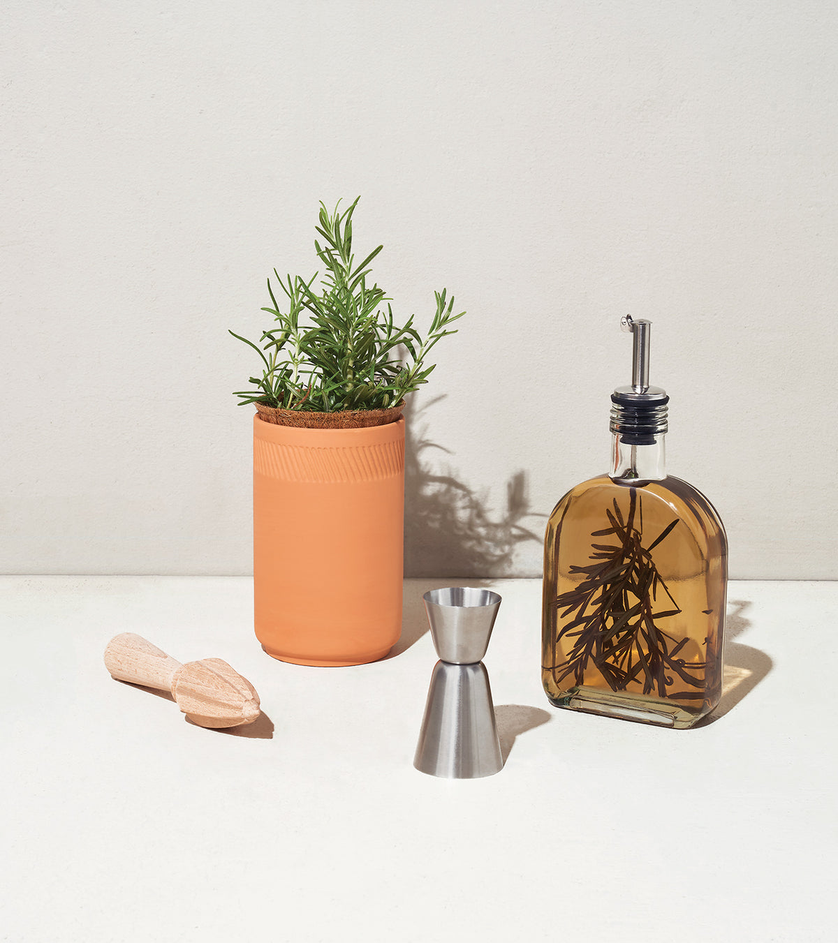 Botanical gift box set featuring a potted rosemary plant, infused oil, a jigger, and a wooden muddler on a neutral background.