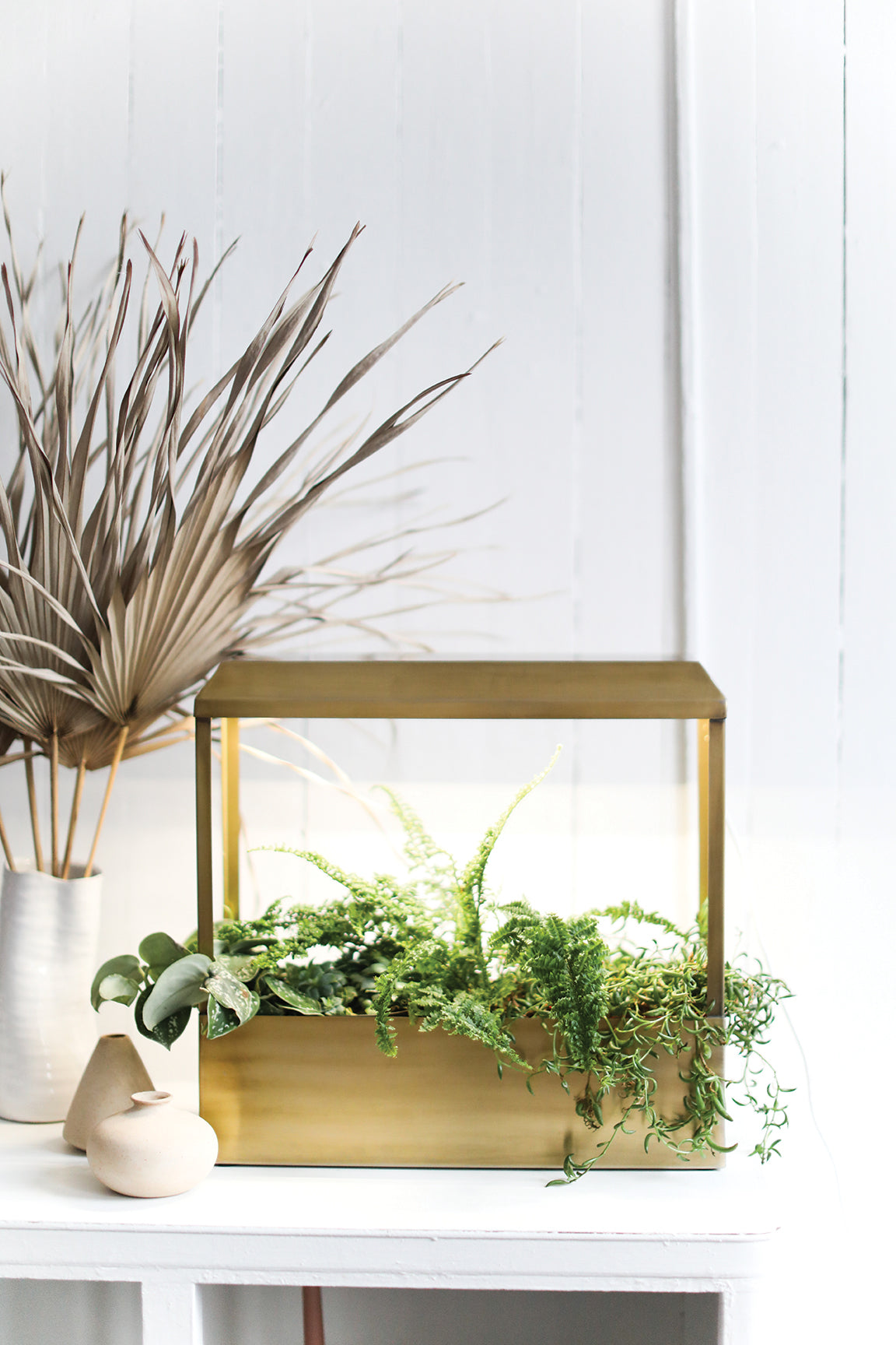 A modern Smart Growhouse with a brass frame, featuring lush green plants, displayed on a white table alongside decorative vases.