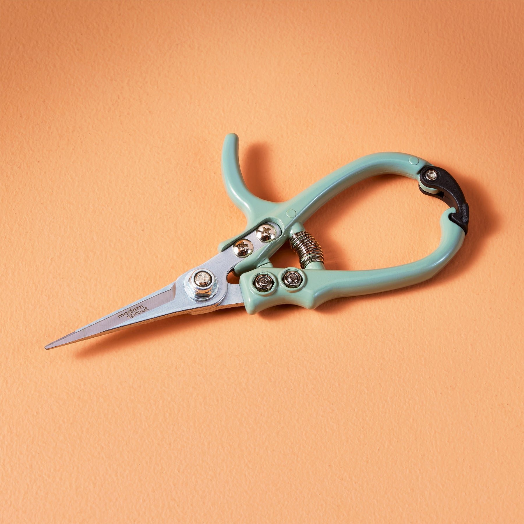 Mint green gardening shears with a stainless steel blade and ergonomic handle, resting on an orange background.