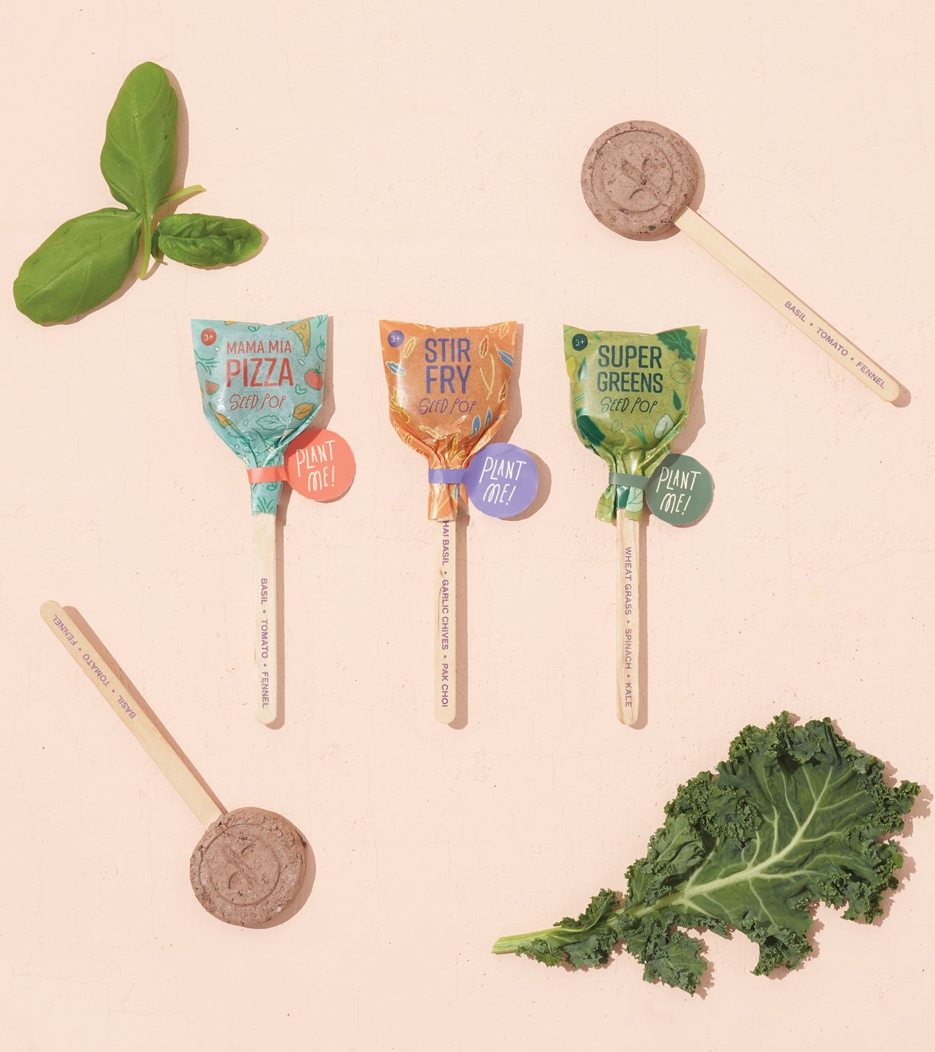Three Seed Lollipops labeled Mamma Mia Pizza, Stir Fry, and Super Greens arranged with fresh basil and kale on a light background.