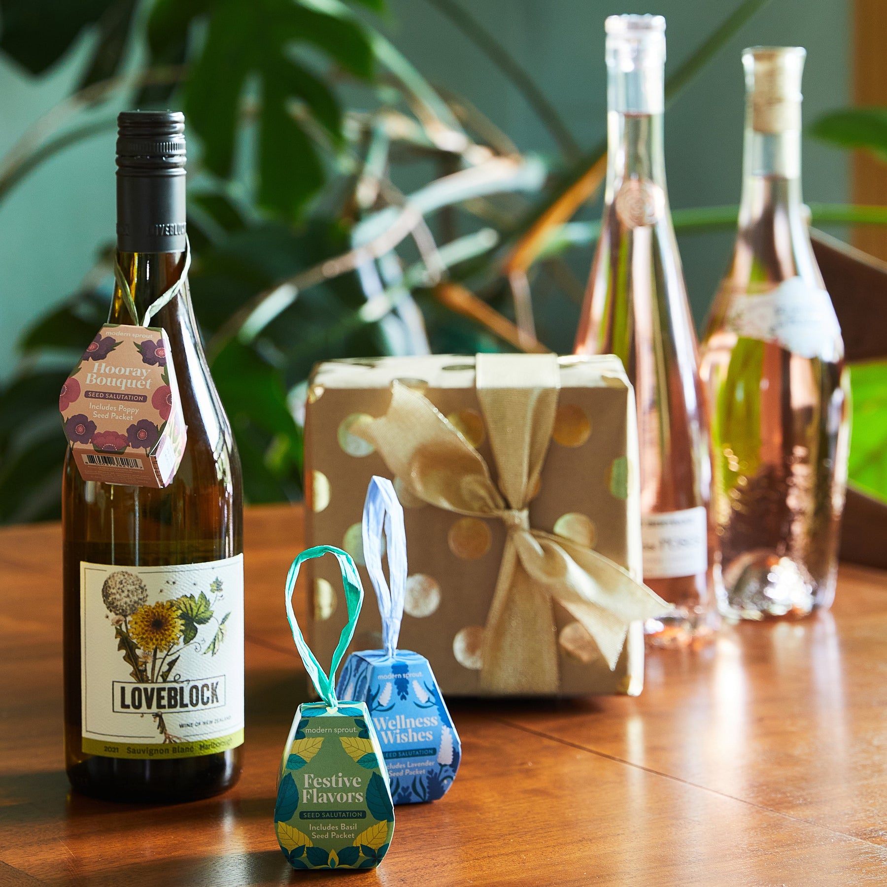 A wooden table displays a wrapped gift with a bow, wine bottles, and seed packets labeled "Festive Flavors" and "Wellness Wishes.