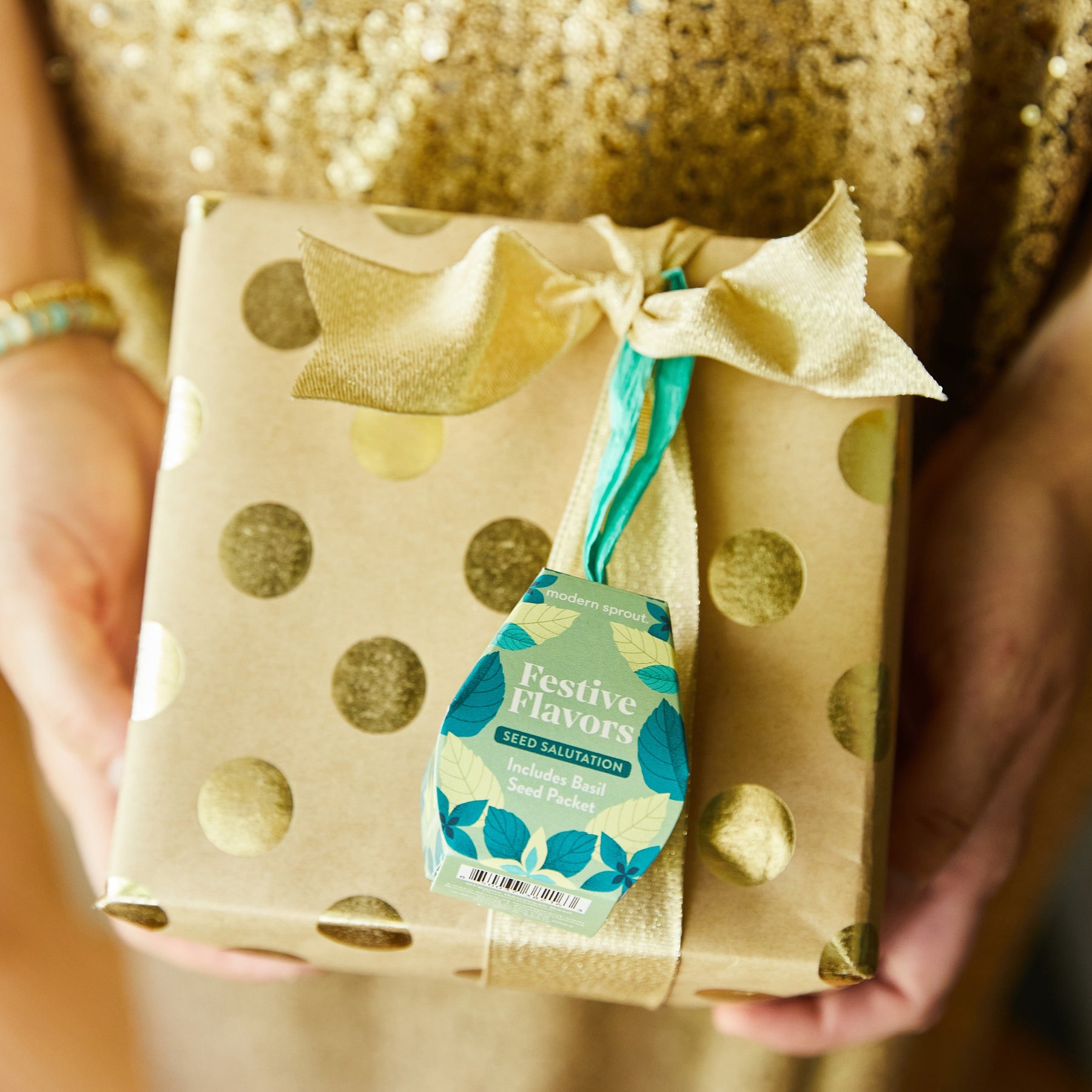 A person holds a beautifully wrapped gift box with gold polka dots, featuring a tag that reads "Festive Flavors" and includes basil seed packets.