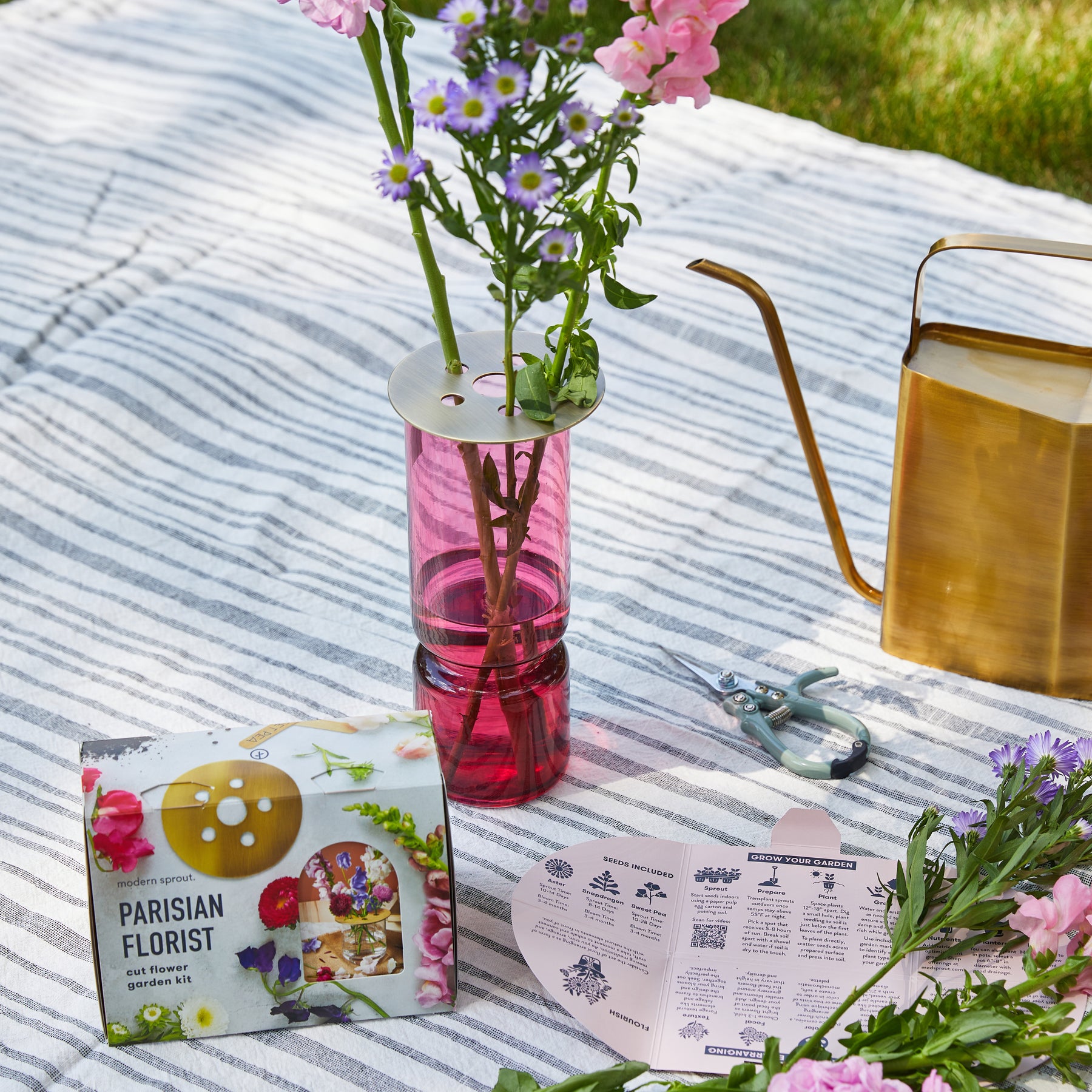 Seed collection box with floral accessories, a pink vase with flowers, watering can, and gardening tools on a striped blanket.