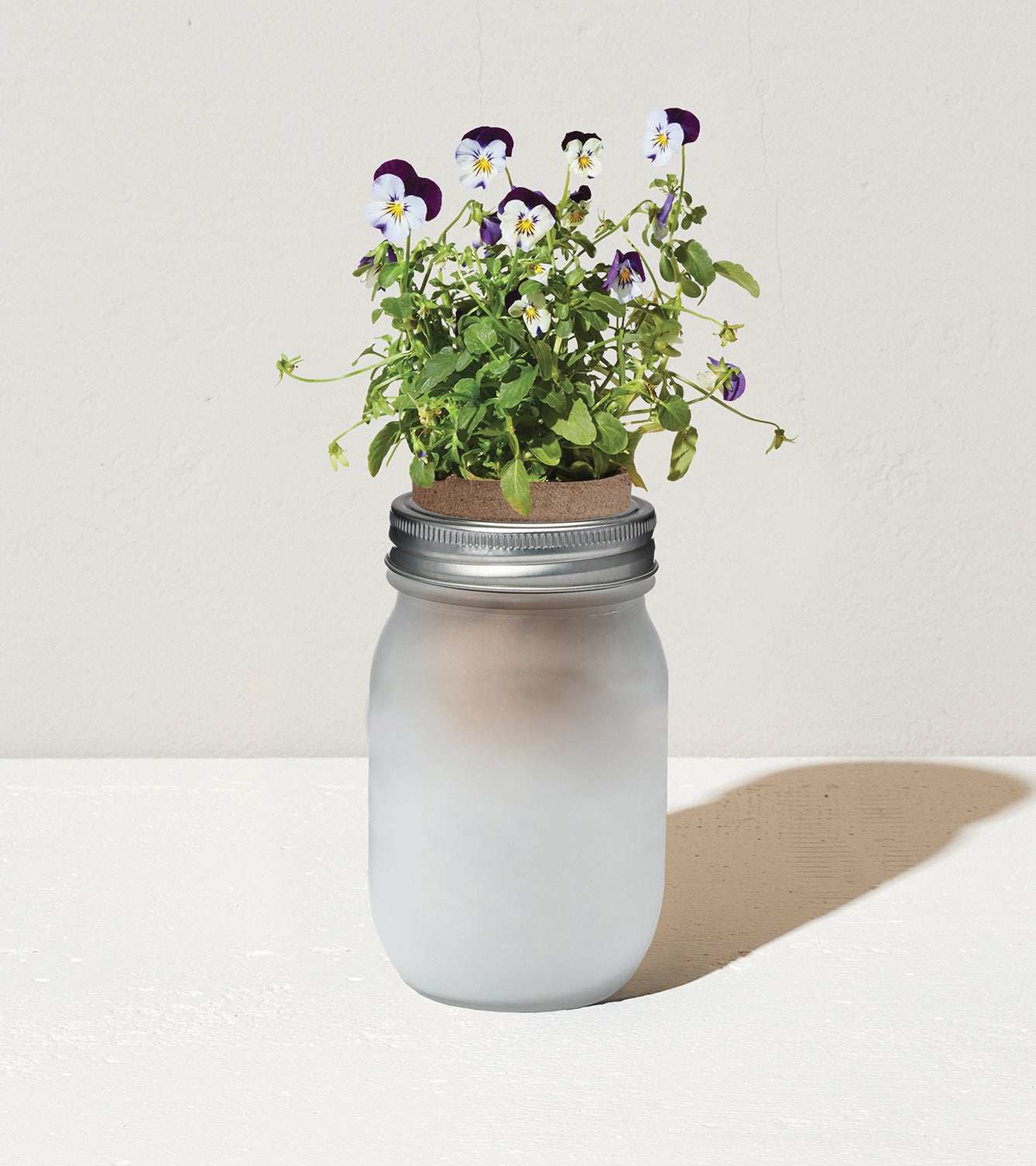 Frosted glass jar with a metal lid, featuring vibrant purple and white flowers and green foliage on top.