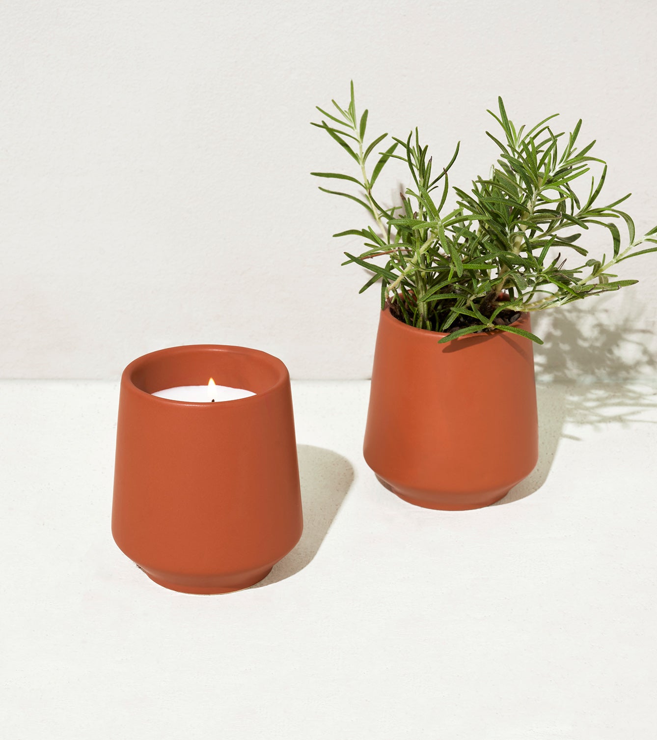 Two terracotta-colored vessels on a light surface: one holds a lit candle, and the other contains fresh rosemary.