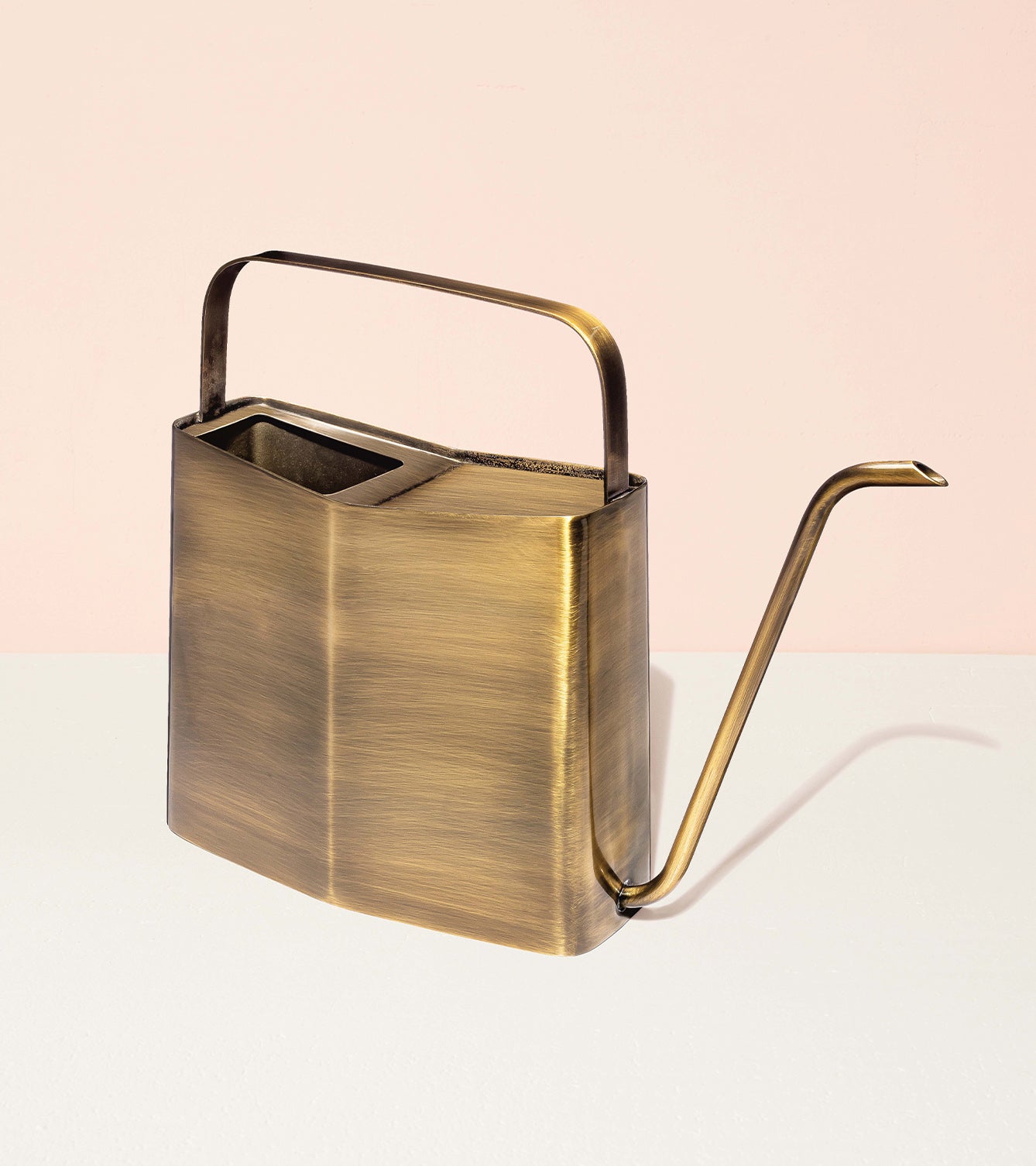Brass watering can with a sleek design and long spout, set against a soft pink background.