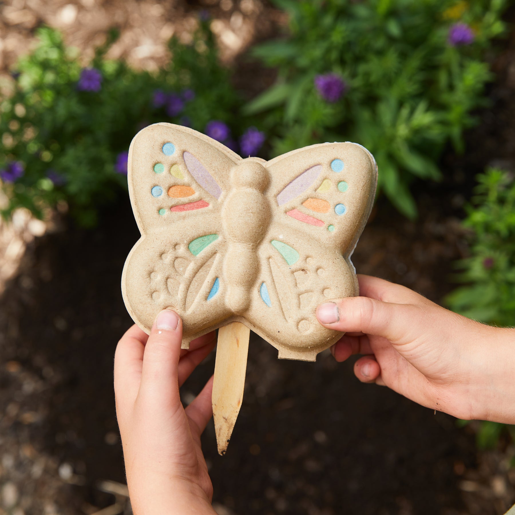 Child holding a colorful butterfly-shaped garden kit in a garden setting, surrounded by green plants and flowers.