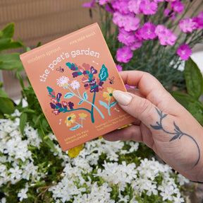 Hand holding a colorful package of Bespoke Blooms Seed Balls titled "The Poet's Garden," surrounded by blooming flowers.